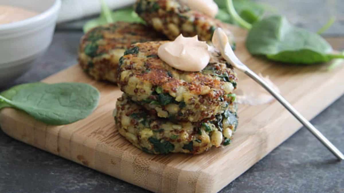 Three quinoa and spinach patties stacked on a wooden board, topped with a dollop of sauce and garnished with spinach leaves. A spoon rests beside the stack.