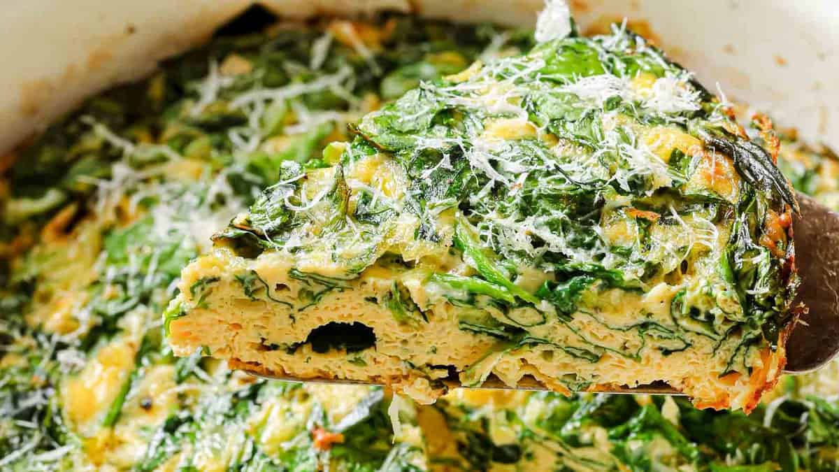 A close-up of a spinach and cheese frittata slice being lifted from a baking dish, topped with grated cheese.