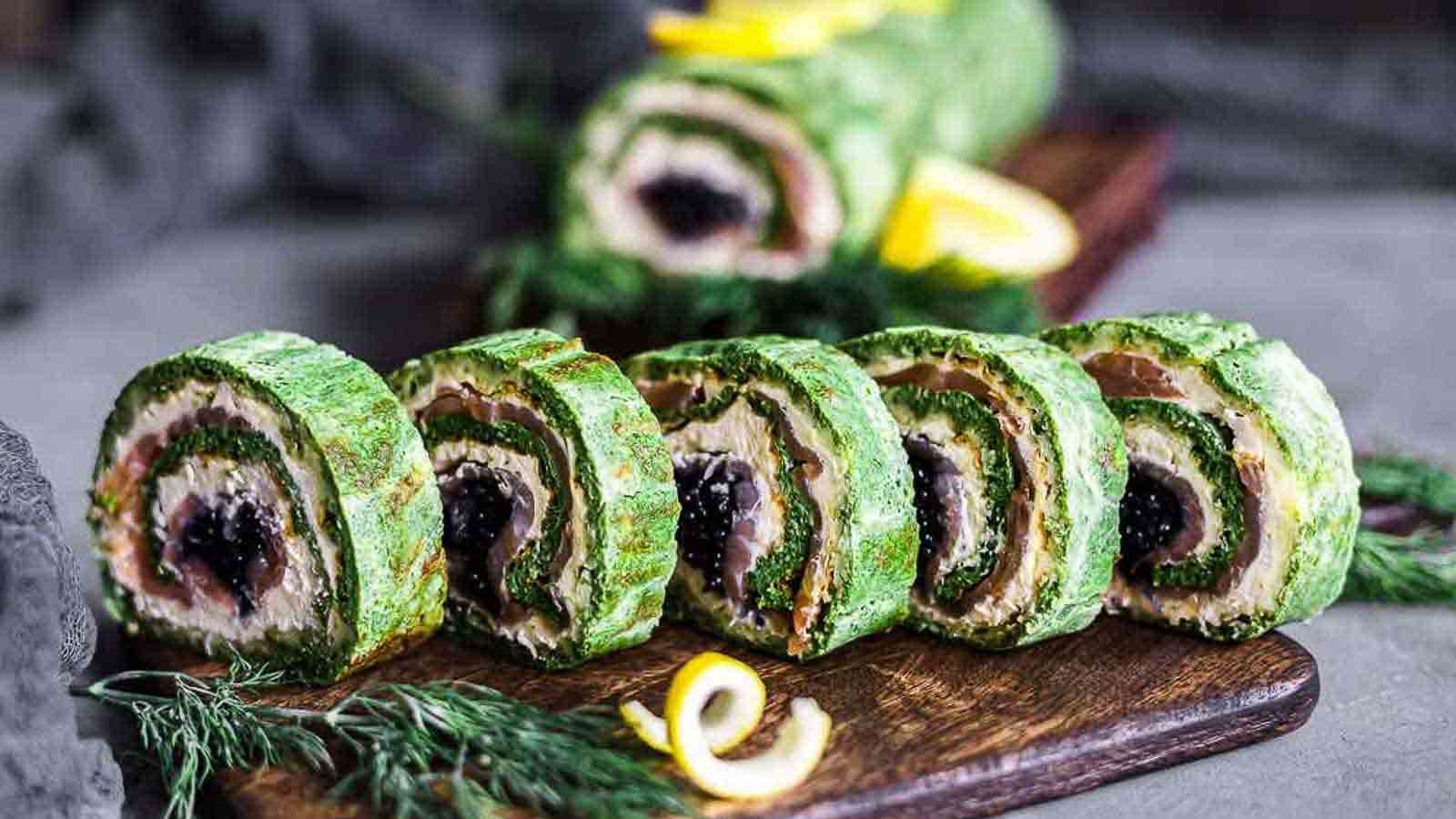 Sliced green roulade with visible layers of cream and dark filling, arranged neatly on a wooden board with garnish and lemon slices.