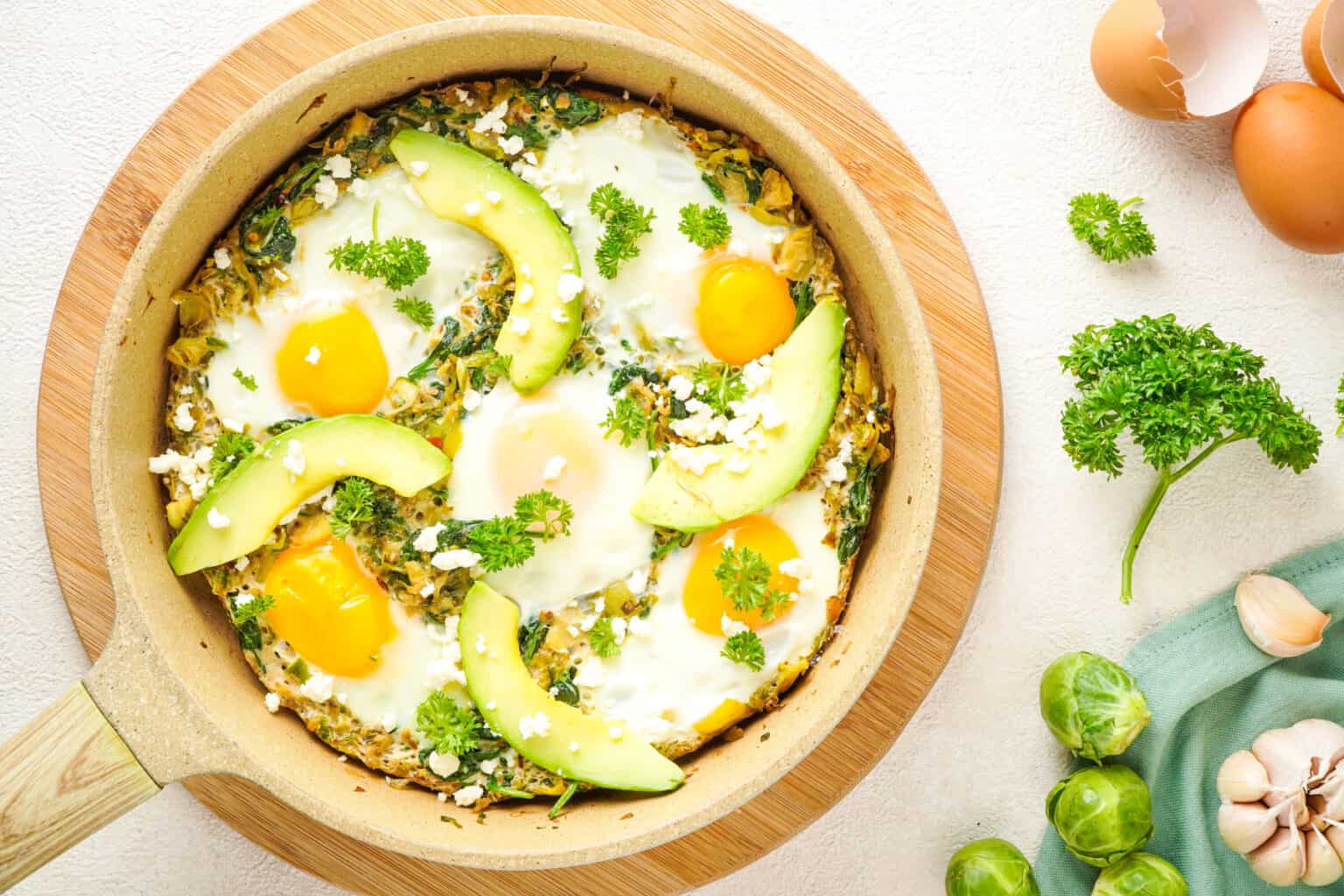 A skillet with baked eggs, avocado slices, crumbled cheese, and parsley garnish, surrounded by eggs, garlic, parsley, and Brussels sprouts on a light surface.