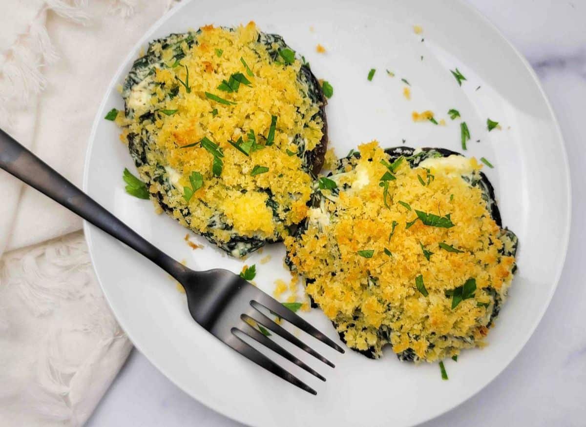 Two baked stuffed portobello mushrooms topped with golden breadcrumbs and chopped parsley on a white plate with a black fork.