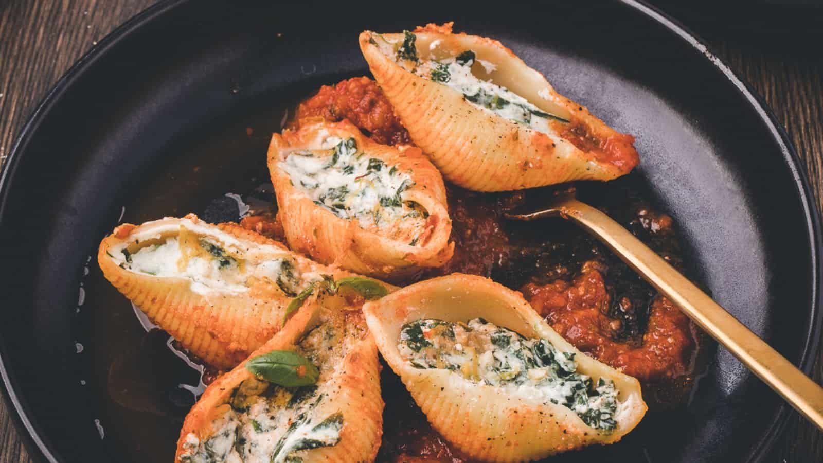 Five stuffed pasta shells filled with ricotta and spinach are served on a black plate with tomato sauce and a gold fork.