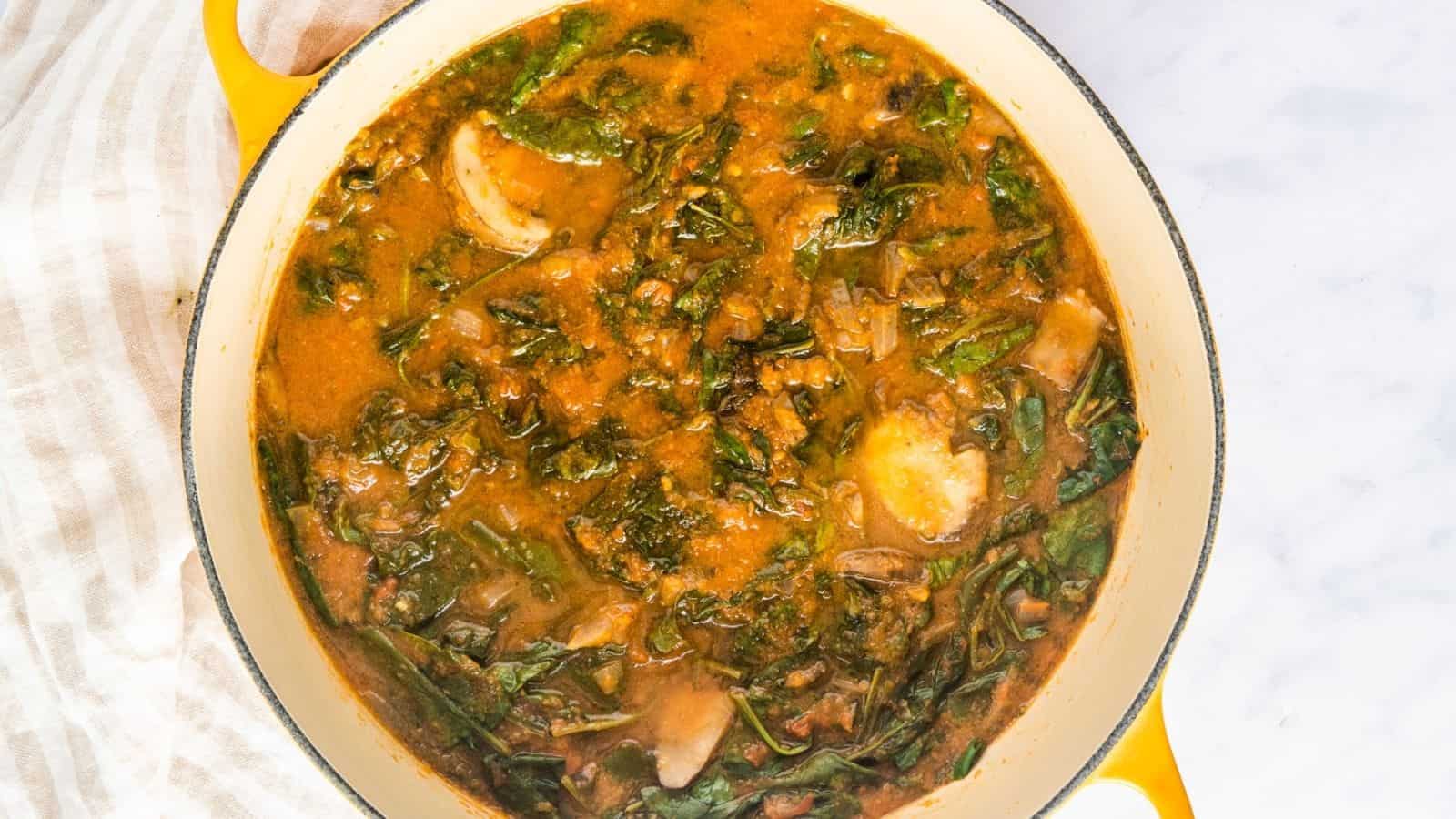 A pot of tomato-based stew with leafy greens and sliced mushrooms on a marble surface.
