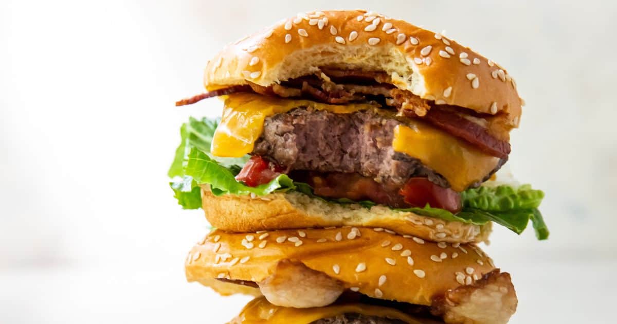 A cheeseburger with a sesame seed bun, lettuce, tomato, bacon, cheddar cheese, and a bite taken out of it.