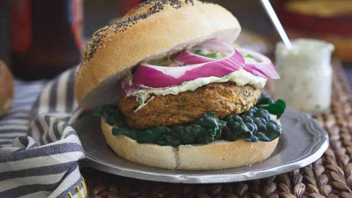 A veggie burger topped with leafy greens, a creamy spread, and sliced red onions on a poppy seed bun sits on a plate.