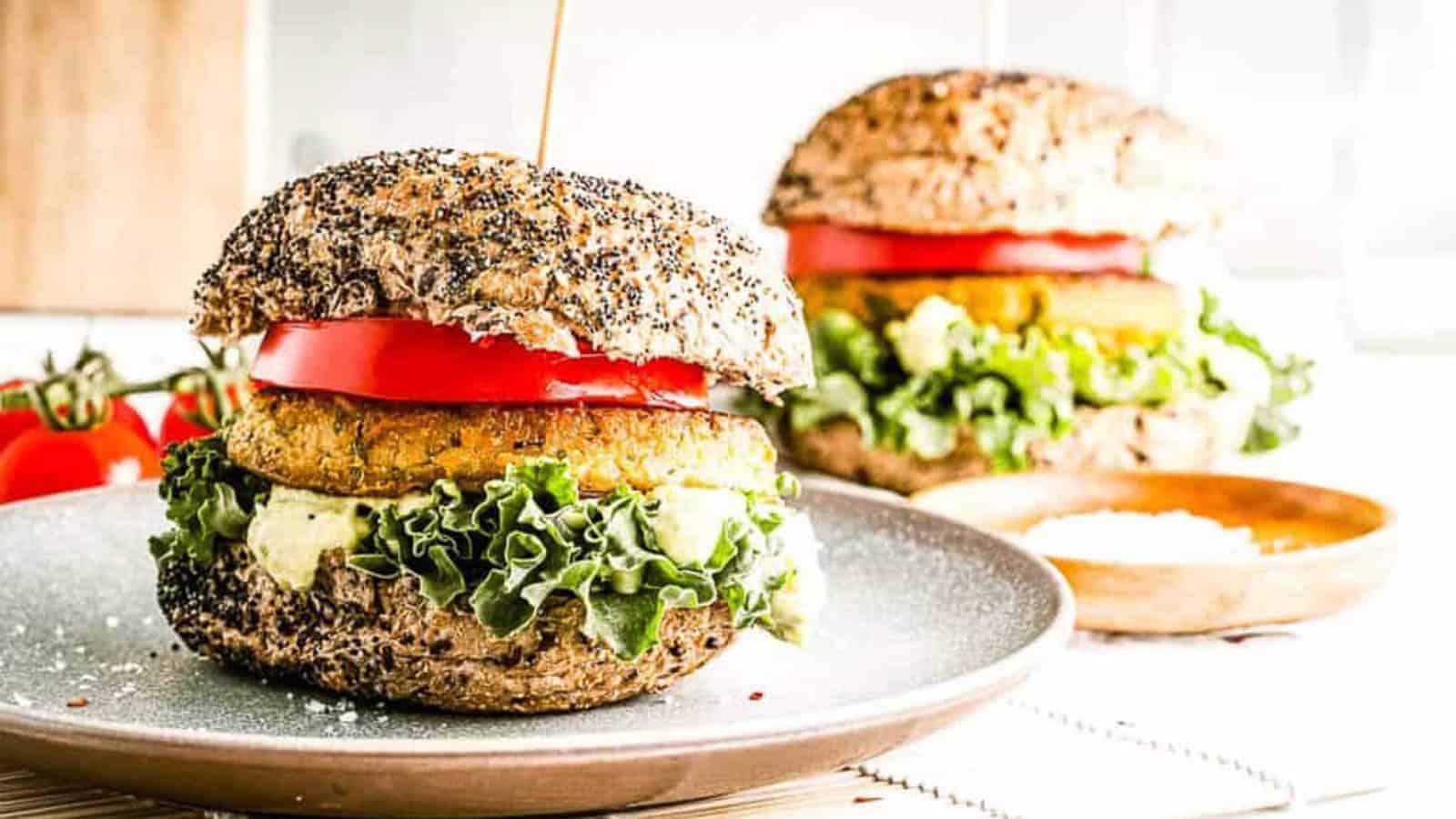 A veggie burger with leafy lettuce, tomato slices, and sauce on a seeded whole grain bun sits on a plate, with a similar burger in the background.