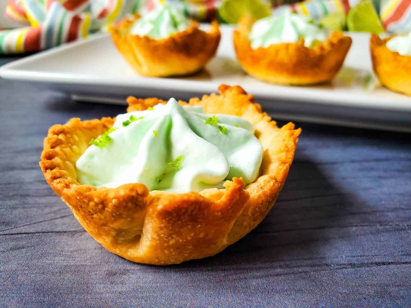 A close-up of a tart with a golden crust filled with whipped cream and garnished with lime zest, with similar tarts on a plate in the background.