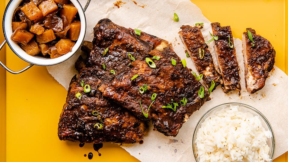 Barbecued ribs sliced on parchment with chopped green onions, served with a bowl of white rice and a bowl of cooked cubed root vegetables on a yellow surface.