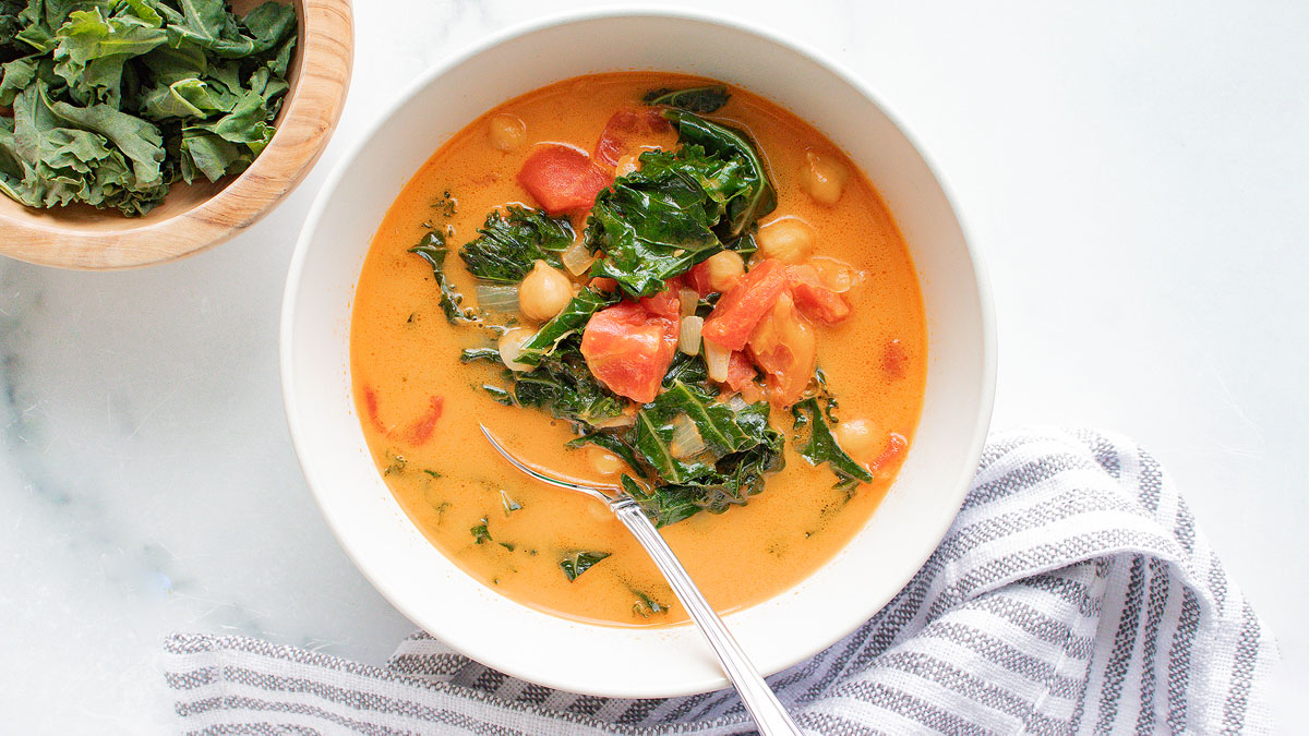 A bowl of vegetable soup with chickpeas, tomatoes, and leafy greens sits on a white surface beside a bowl of raw greens and a striped cloth.