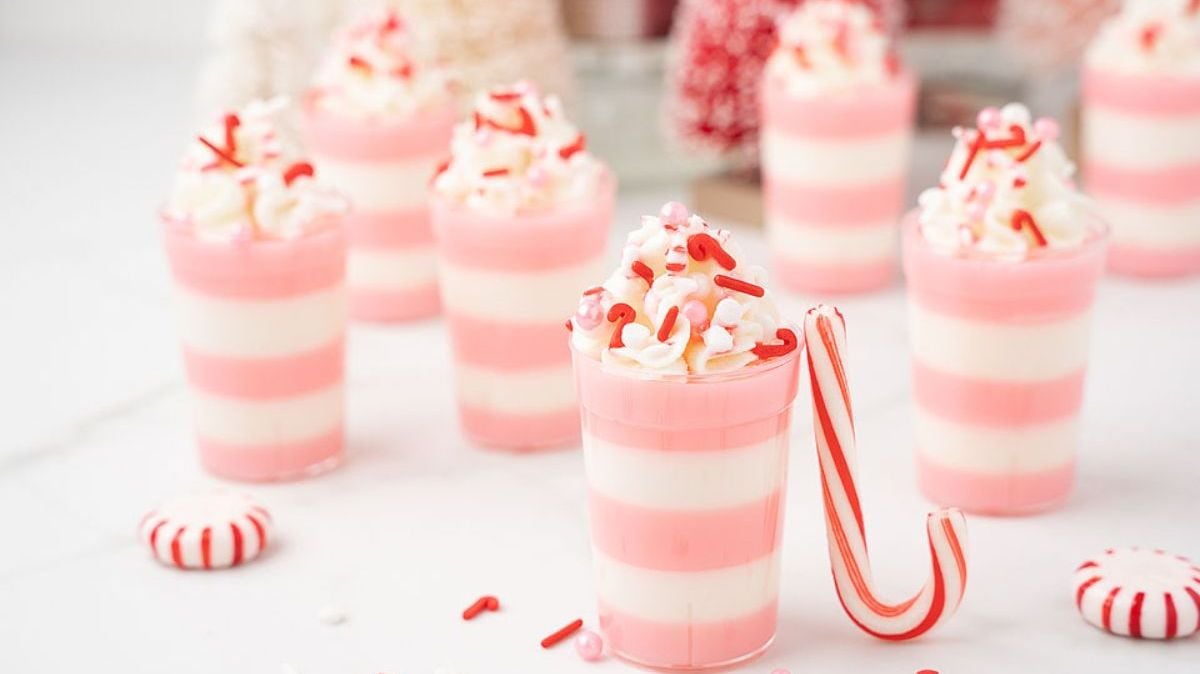 Several pink and white striped dessert cups topped with whipped cream and red sprinkles are arranged on a white surface with a mini candy cane and peppermint candies nearby.