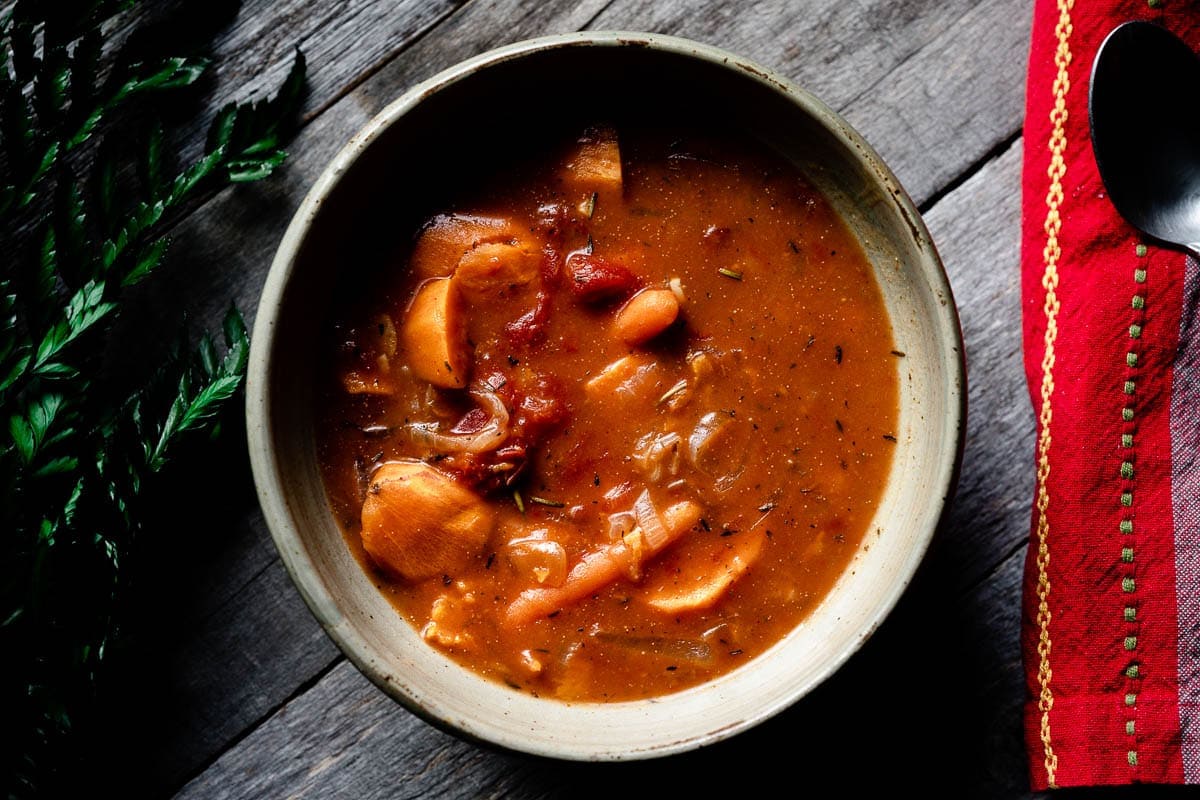 A bowl of vegetable soup with chunks of sweet potato and tomatoes, placed on a wooden surface next to a red napkin and a spoon.
