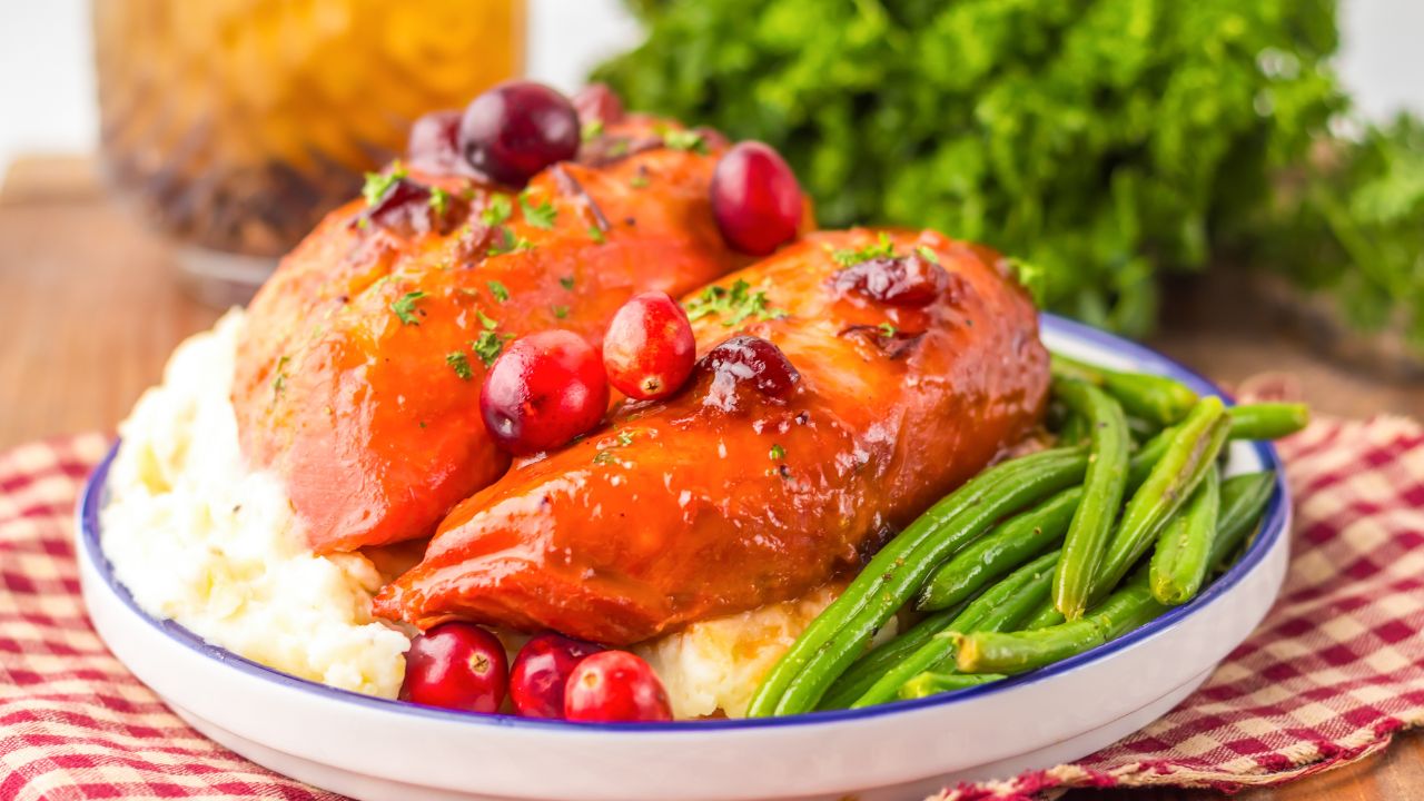 A plate of glazed chicken breast topped with cranberries, served over mashed potatoes with a side of green beans.