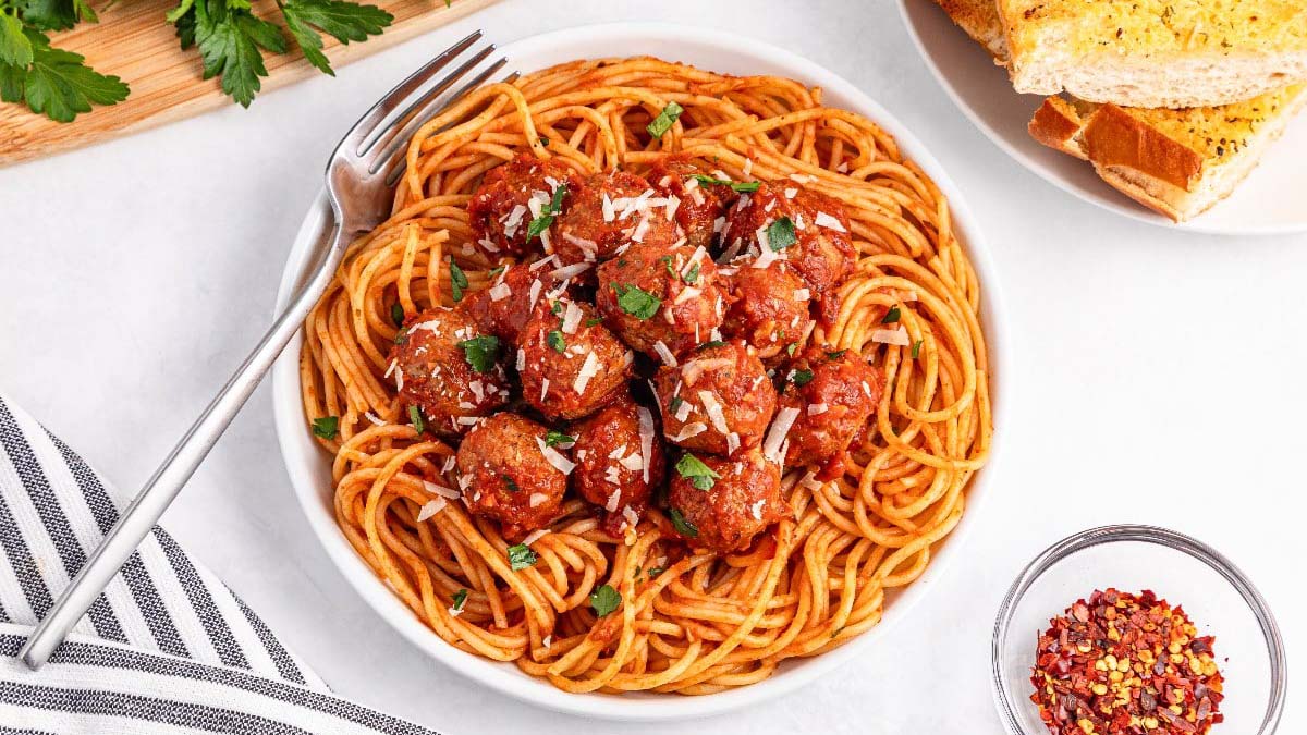 A bowl of spaghetti with meatballs in tomato sauce, topped with grated cheese and parsley, with a fork, garlic bread, and red pepper flakes on the side.