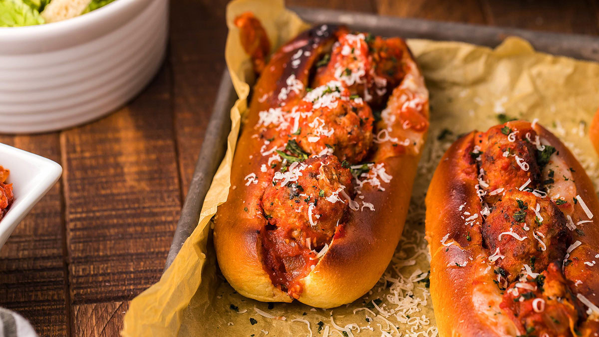 Two toasted sub rolls filled with meatballs in tomato sauce, topped with grated cheese and herbs, sit on parchment paper atop a baking tray.