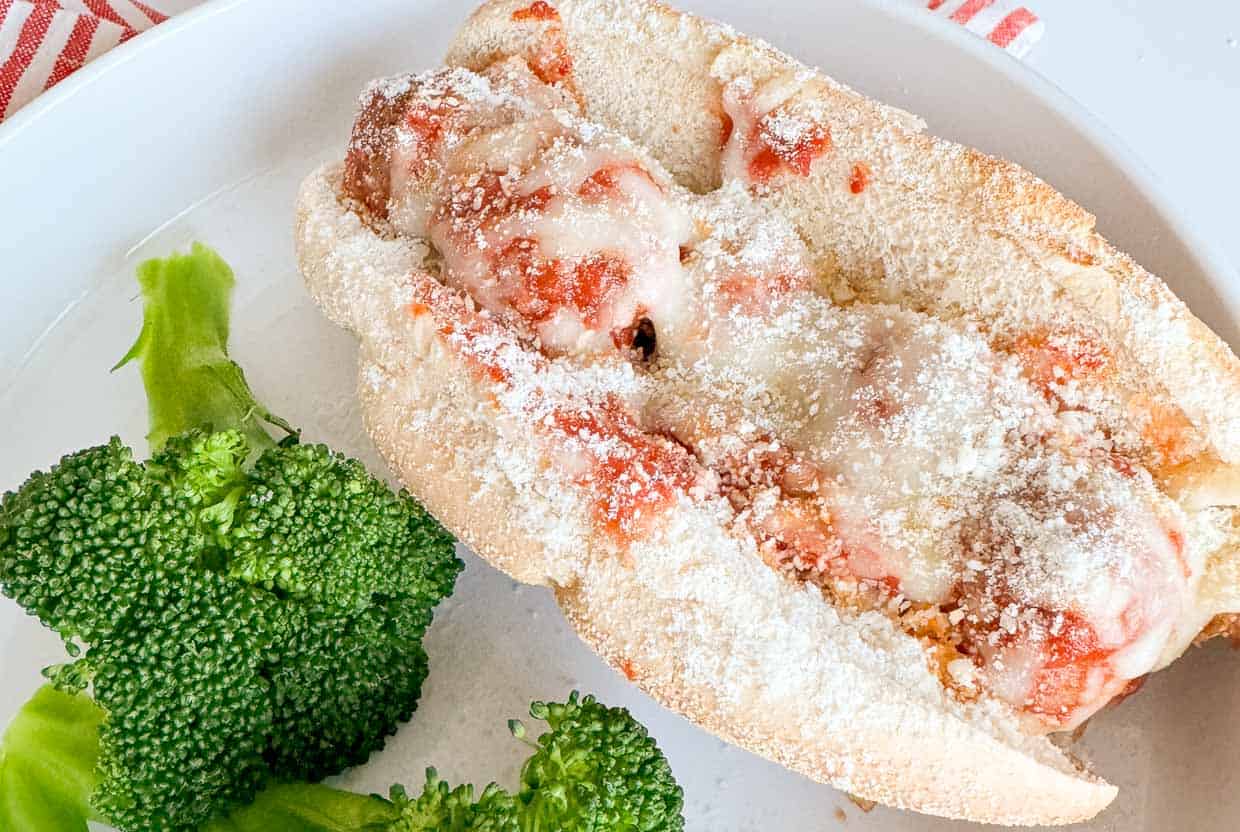 A meatball sub topped with marinara sauce and grated cheese is served on a white plate with a side of steamed broccoli.