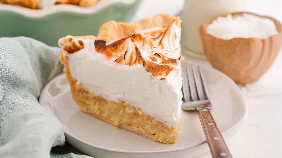 A slice of coconut cream pie with toasted meringue topping on a white plate, next to a fork and a light green napkin.