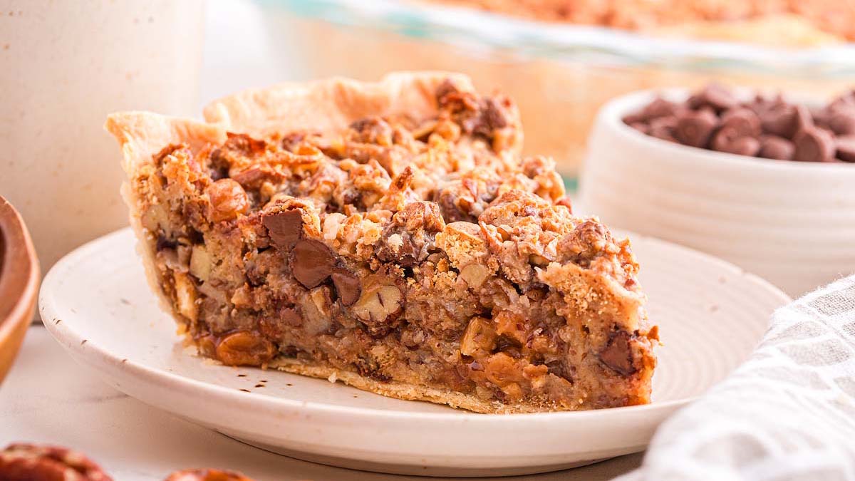 A slice of nut and chocolate chip pie sits on a white plate, with a bowl of chocolate chips and another pie in the background.