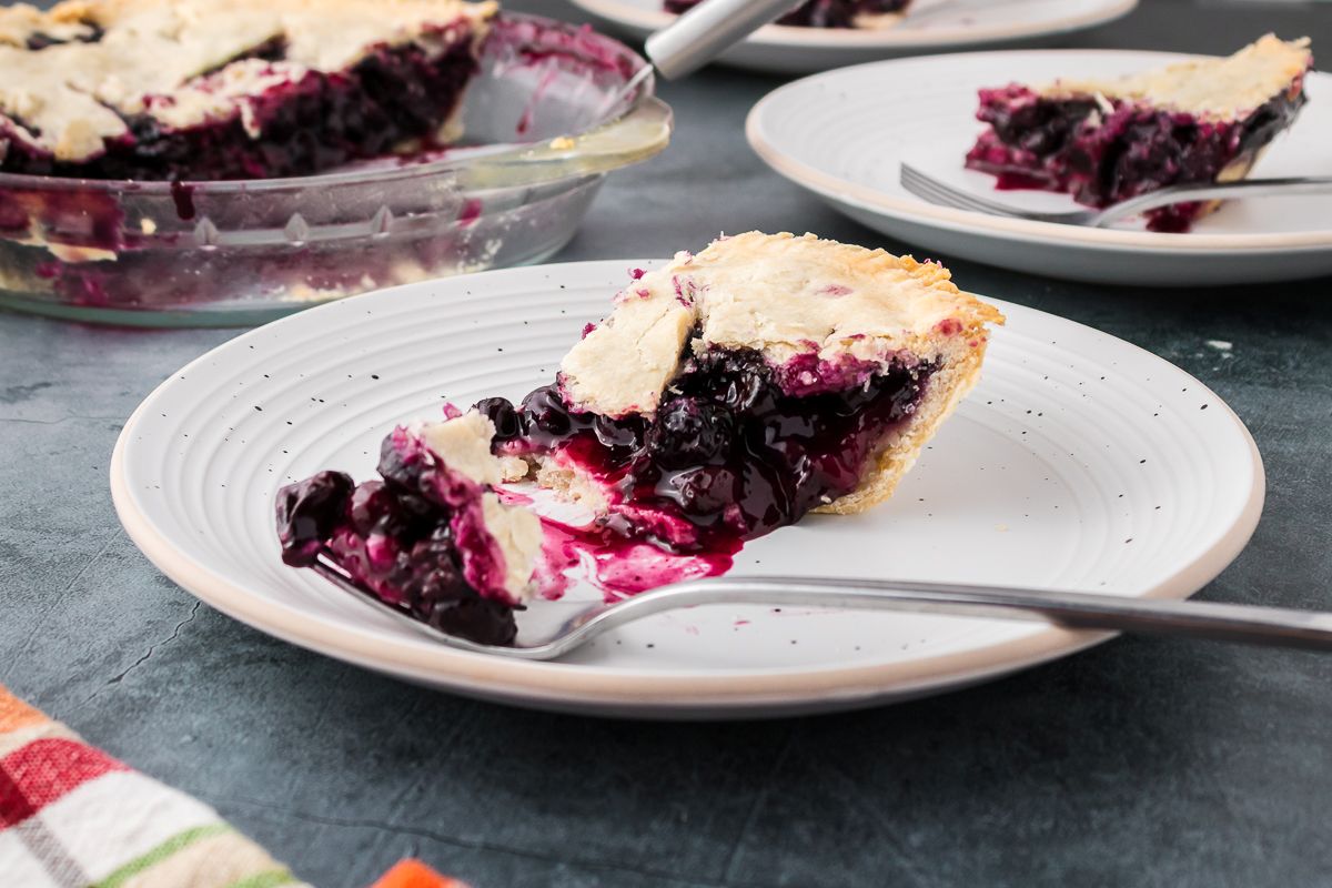A slice of blueberry pie with a flaky crust sits on a white plate, with a fork holding a bite-sized piece; the rest of the pie is in a glass dish in the background.