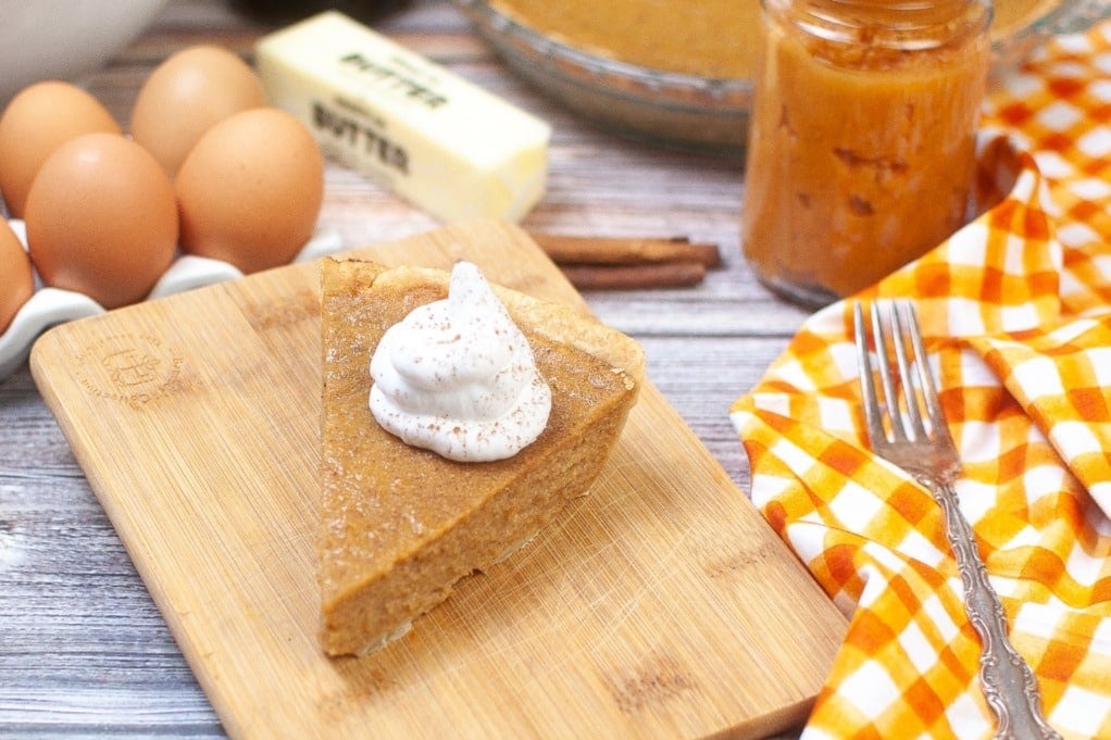 A slice of pumpkin pie with whipped cream on a wooden board, surrounded by eggs, butter, a jar of filling, and an orange checkered napkin with a fork.