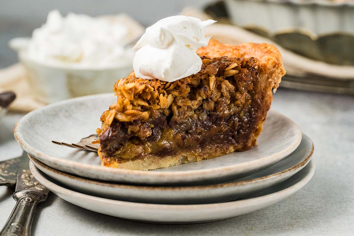 A slice of pie with a golden crust and oat topping, filled with a gooey, dark brown filling, topped with a dollop of whipped cream, served on stacked white plates.