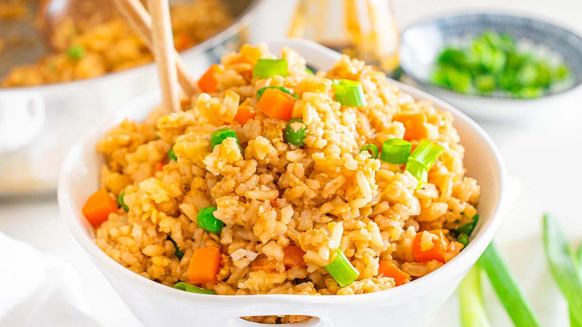 A white bowl filled with fried rice, garnished with chopped green onions and diced carrots, with chopsticks resting inside.