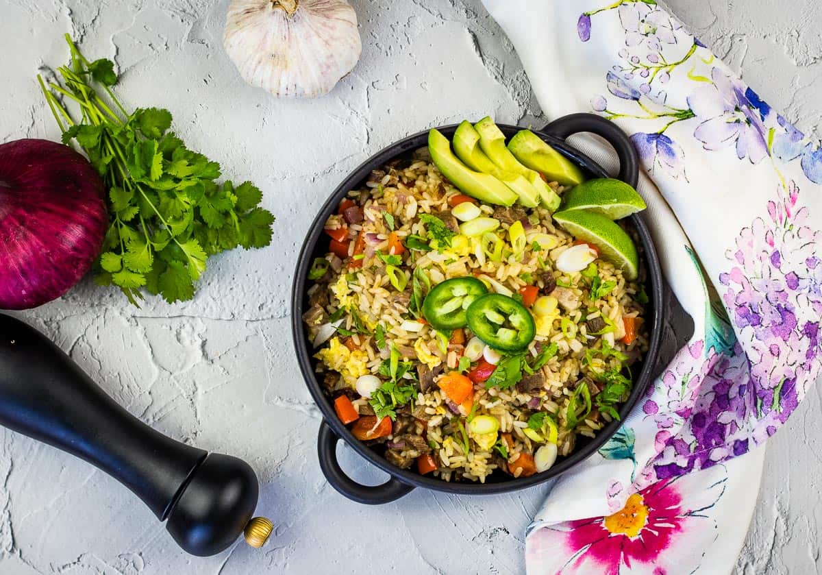 A bowl of vegetable fried rice garnished with lime, avocado, green onions, and jalapeรฑo slices sits on a table with cilantro, red onion, garlic, a pepper grinder, and a floral napkin nearby.