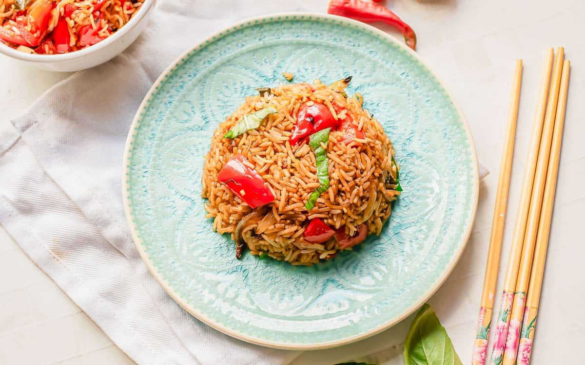 A mound of fried rice with red bell peppers and herbs is served on a light blue plate, with chopsticks beside it on a white table.