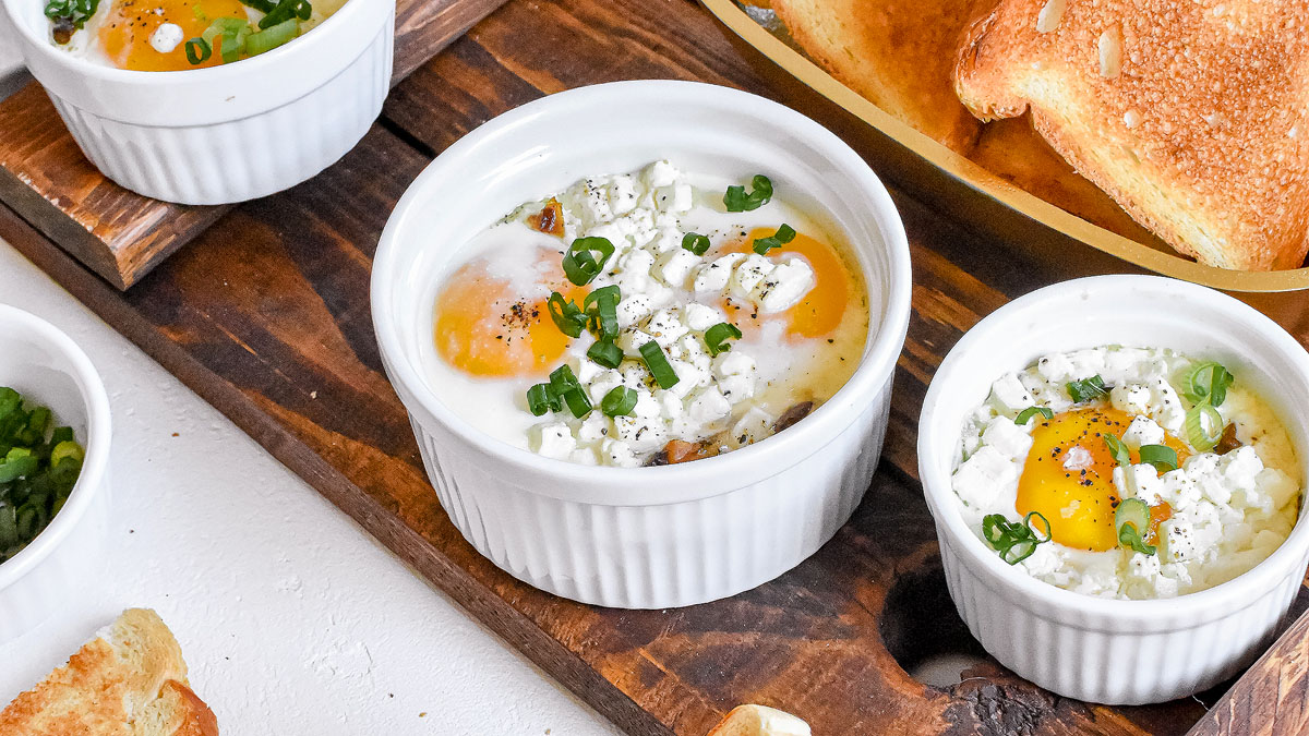 Three white ramekins with baked eggs, crumbled cheese, and chopped green onions on a wooden tray, next to slices of toasted bread.