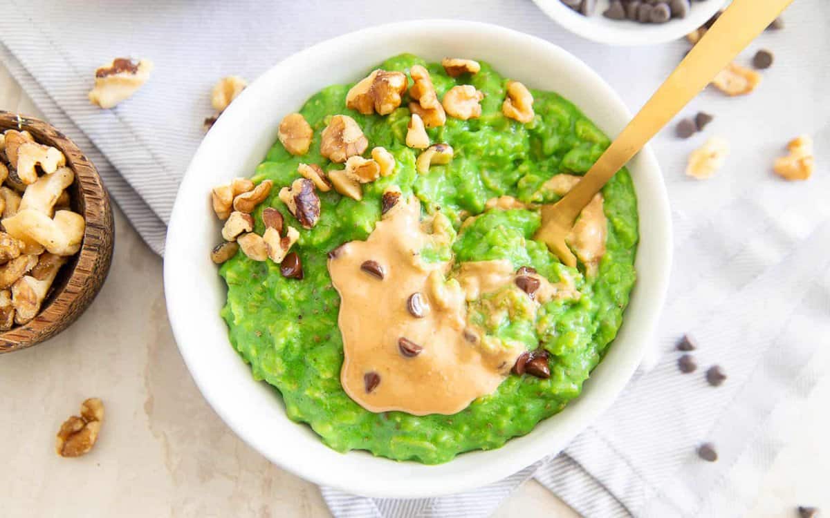 A white bowl filled with bright green oatmeal, topped with walnut pieces, a dollop of peanut butter, and chocolate chips, with a gold spoon resting inside.