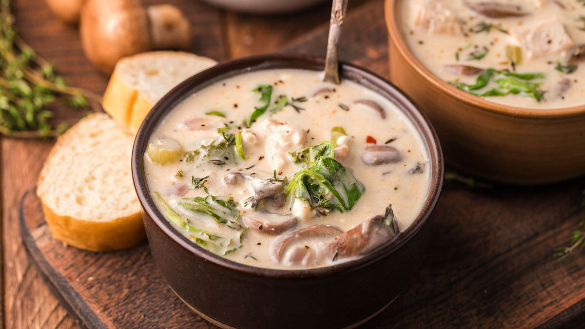 A bowl of creamy soup with mushrooms, greens, and herbs, accompanied by slices of bread on a wooden surface.