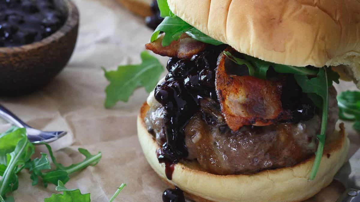 Close-up of a hamburger with bacon, arugula, and a dark sauce, served on a bun. A bowl of sauce and arugula leaves are in the background.