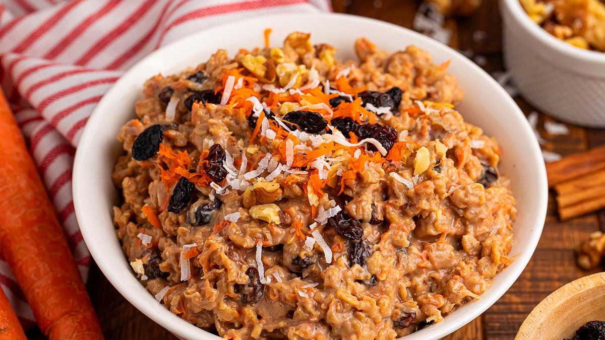A bowl of carrot cake oatmeal topped with shredded coconut, raisins, grated carrots, and chopped walnuts sits on a wooden surface.
