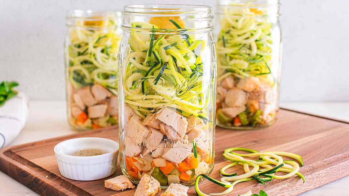 Three mason jars filled with chopped cooked chicken, diced vegetables, and zucchini noodles are arranged on a wooden board next to a small dish of dressing.