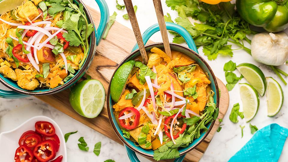 Two bowls of yellow curry with vegetables and herbs, topped with sliced red chili, onions, cilantro, and lime wedges, on a wooden board with fresh ingredients around.