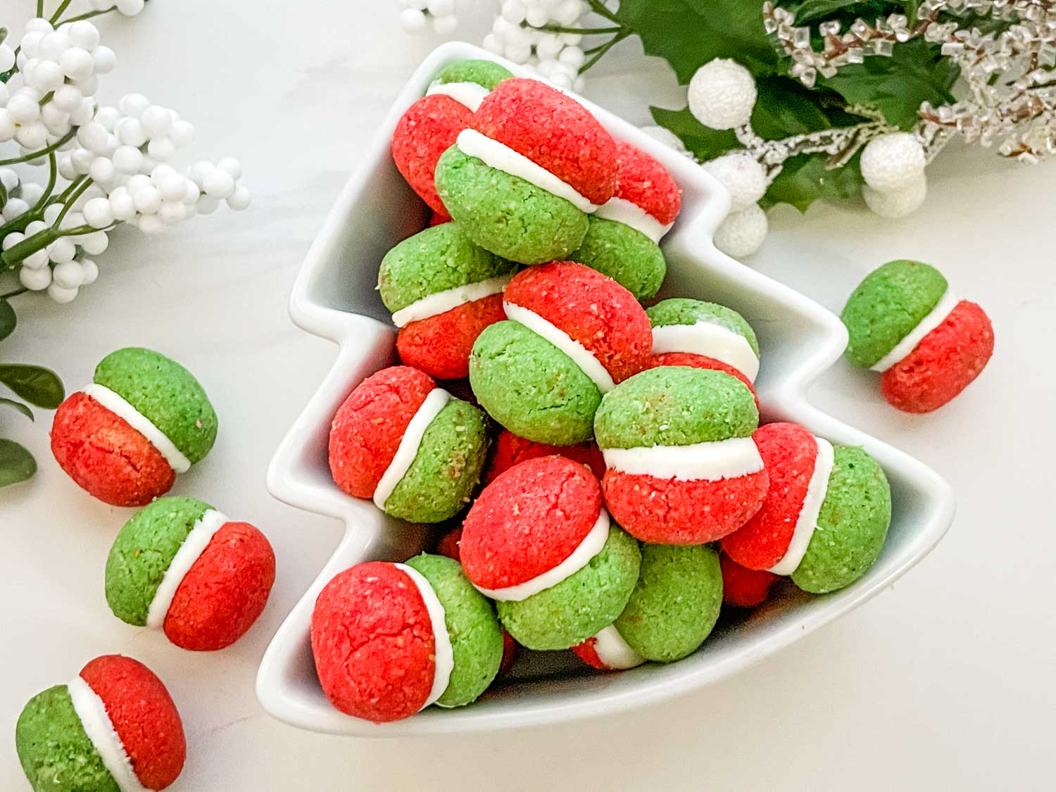 A white tree-shaped dish filled with red and green sandwich cookies with white filling, surrounded by festive holiday decorations.
