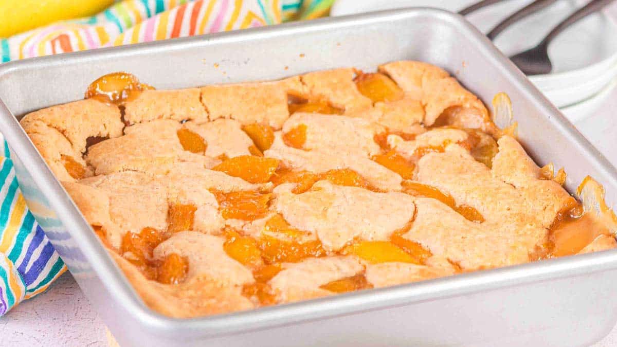 A baked peach cobbler with a golden-brown crust sits in a square metal baking pan on a table.