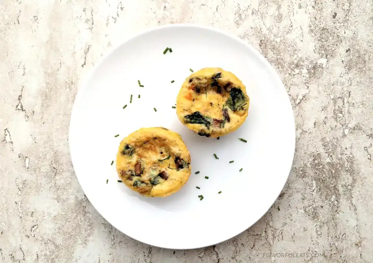 Two baked egg muffins with spinach and vegetables are placed on a round white plate, with a few chopped herbs scattered around.