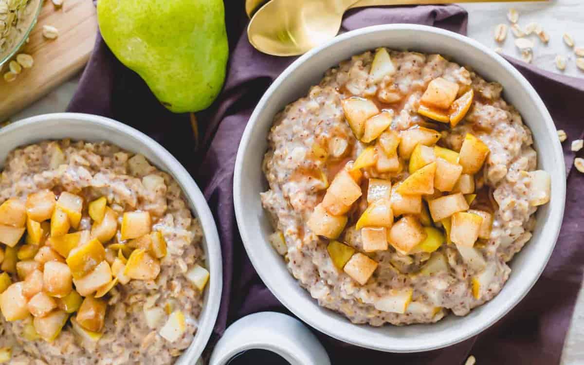Two bowls of oatmeal topped with diced caramelized pears, placed on a dark cloth with a fresh pear, spoon, and oats nearby.