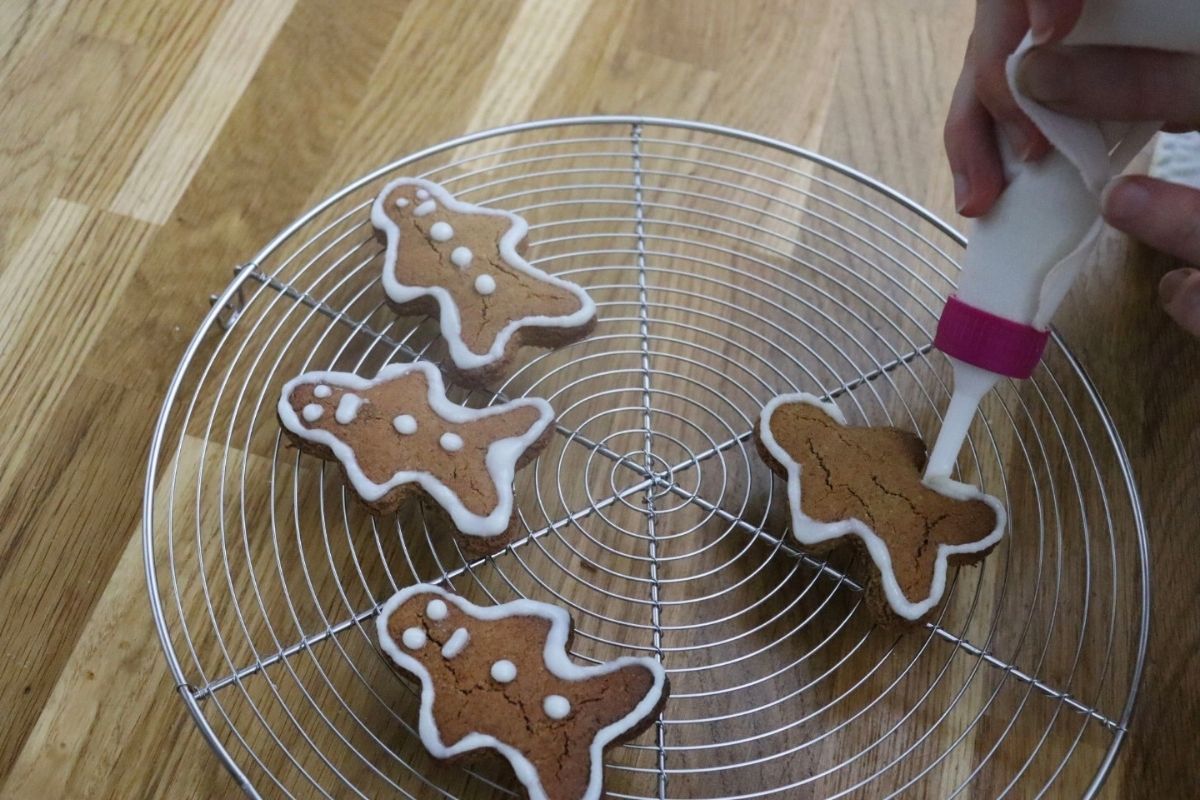 A person decorates gingerbread cookies shaped like people with white icing on a round cooling rack placed on a wooden surface.