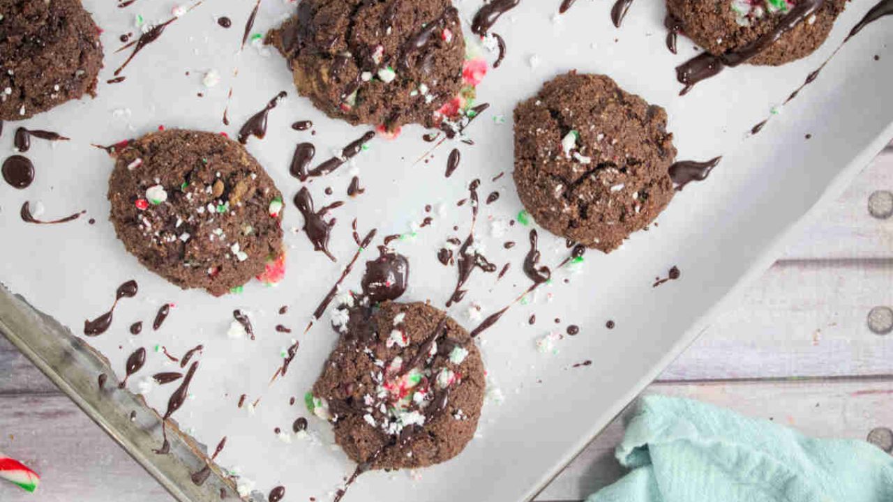 Chocolate cookies with crushed peppermint on top, drizzled with chocolate, arranged on a parchment-lined baking sheet.