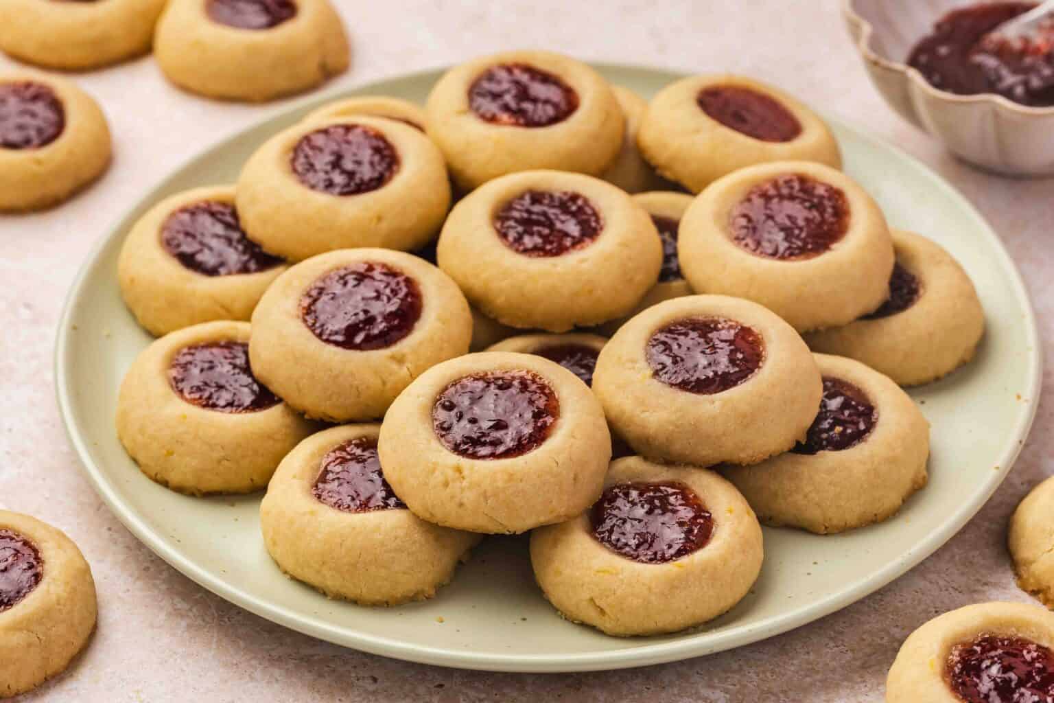 A plate of round thumbprint cookies with jam centers, arranged in a pile, with a few more cookies scattered on the table.
