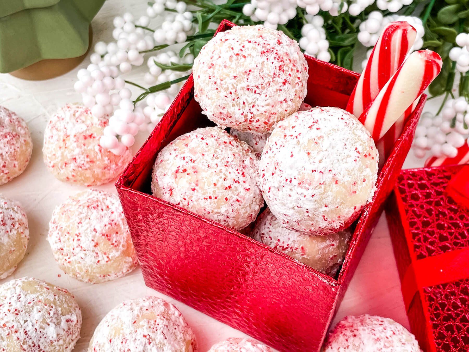 A red gift box filled with powdered sugar-covered cookies and two candy canes, surrounded by more cookies and white artificial berries.