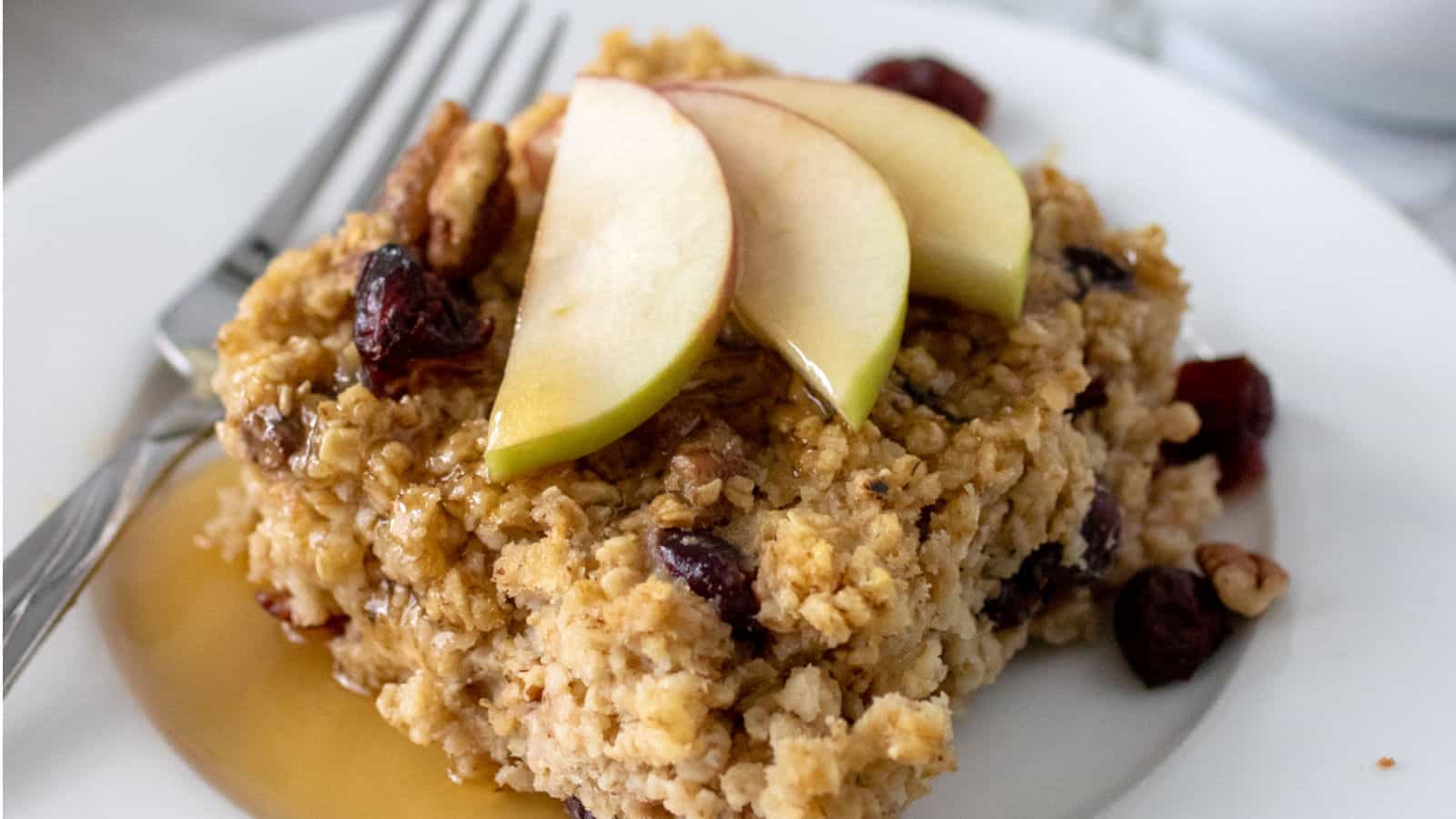 A serving of baked oatmeal topped with apple slices, nuts, and dried cranberries, drizzled with syrup, on a white plate with a fork.