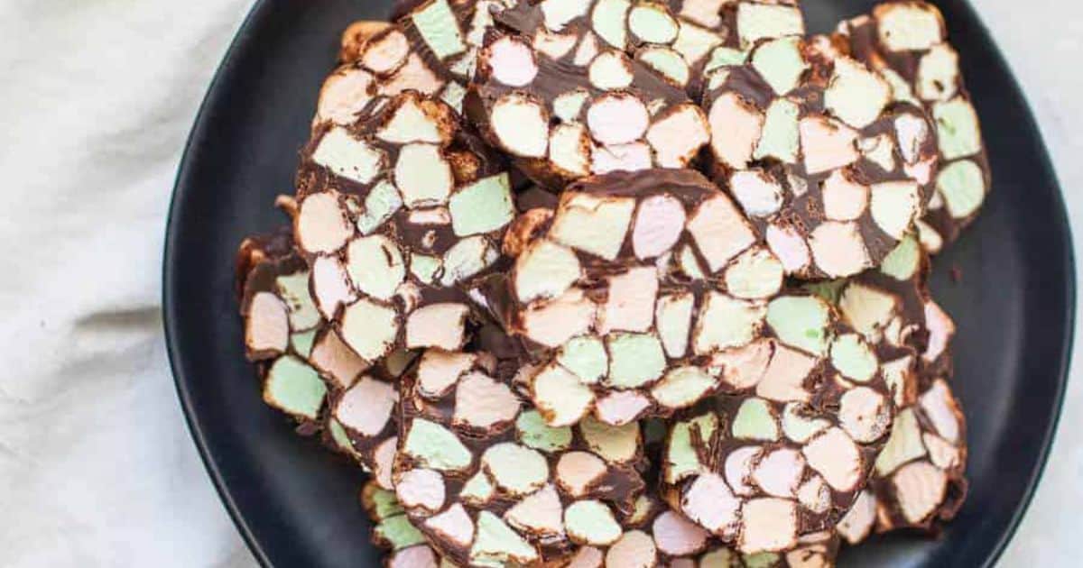 A black plate holds slices of dessert made with pastel-colored mini marshmallows and chocolate, arranged in a stack on a light surface.