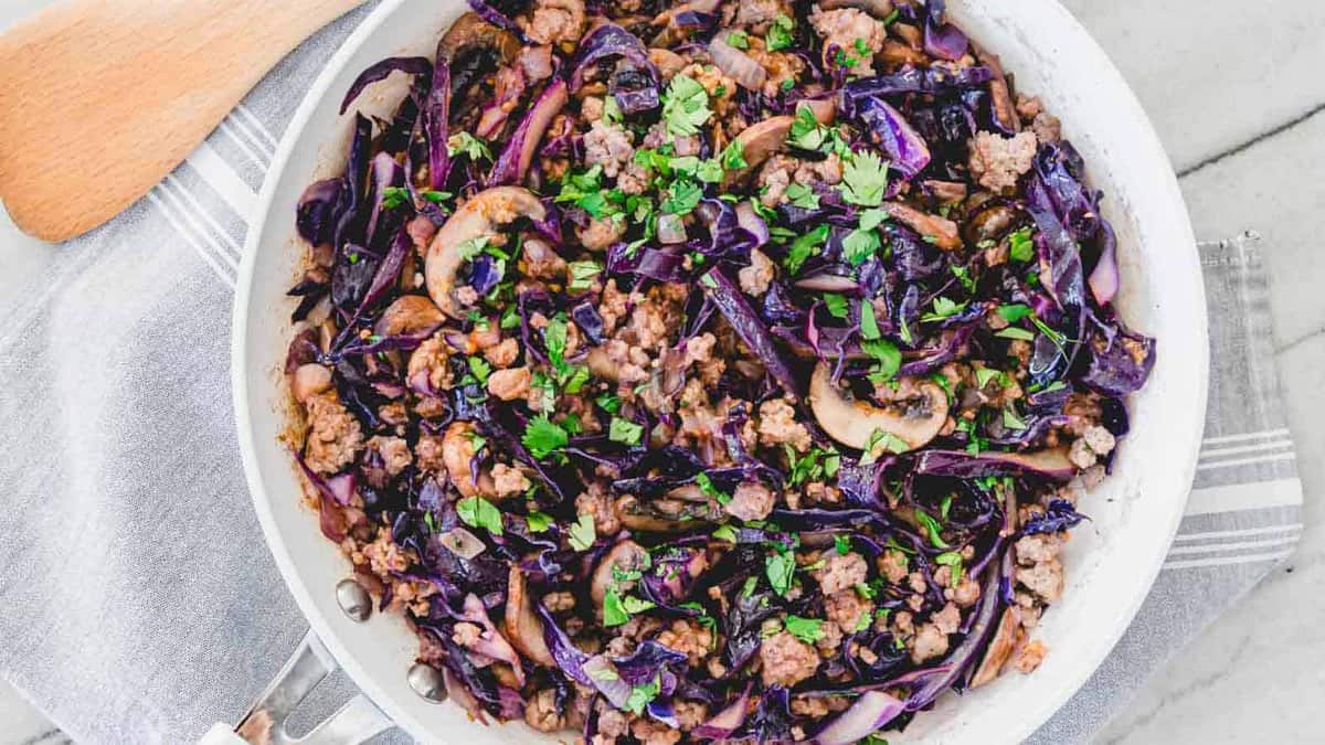 A white skillet filled with cooked ground meat, sliced purple cabbage, mushrooms, and chopped fresh herbs on a kitchen towel.
