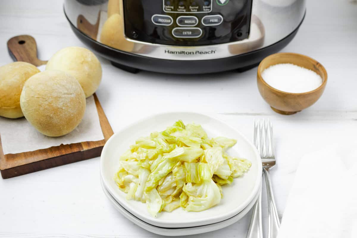 A plate of cooked cabbage sits next to a bowl of salt, three bread rolls on a cutting board, and a slow cooker on a white table.