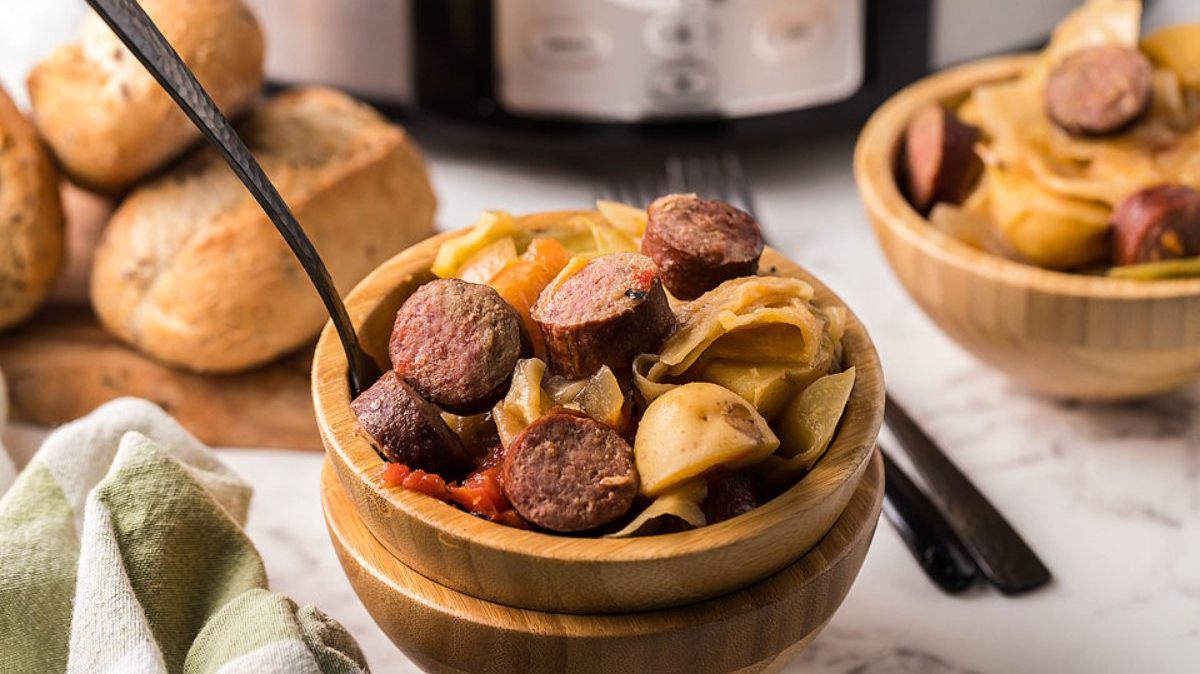 A wooden bowl filled with sliced sausage, potatoes, and vegetables sits on a marble surface with bread rolls and a slow cooker in the background.