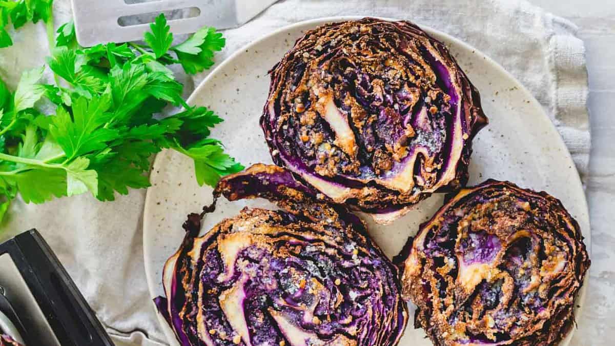 Three roasted purple cabbage steaks on a plate, garnished with fresh parsley on the side and a metal spatula nearby.
