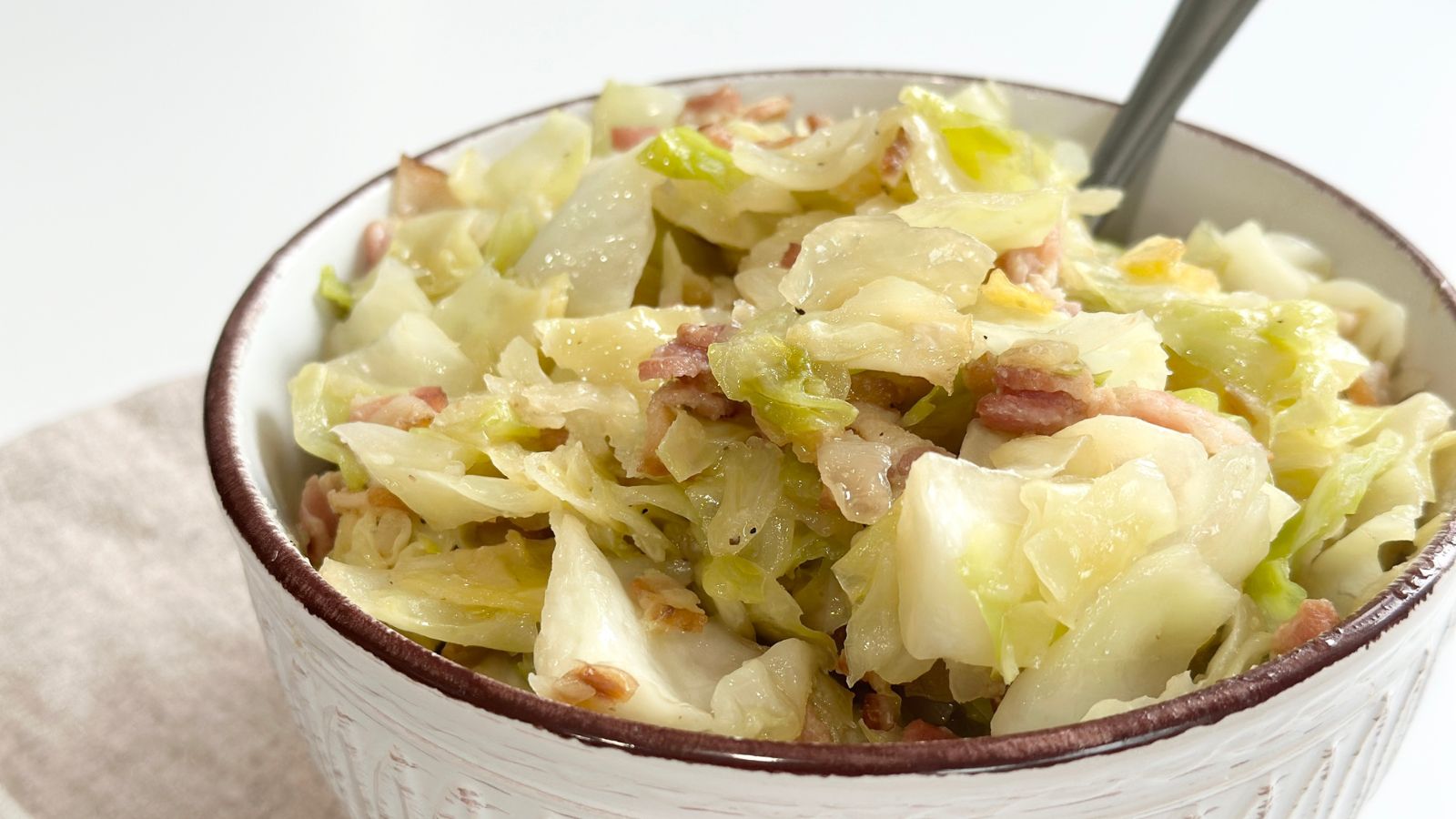 A bowl of cooked cabbage mixed with pieces of bacon, served with a spoon.