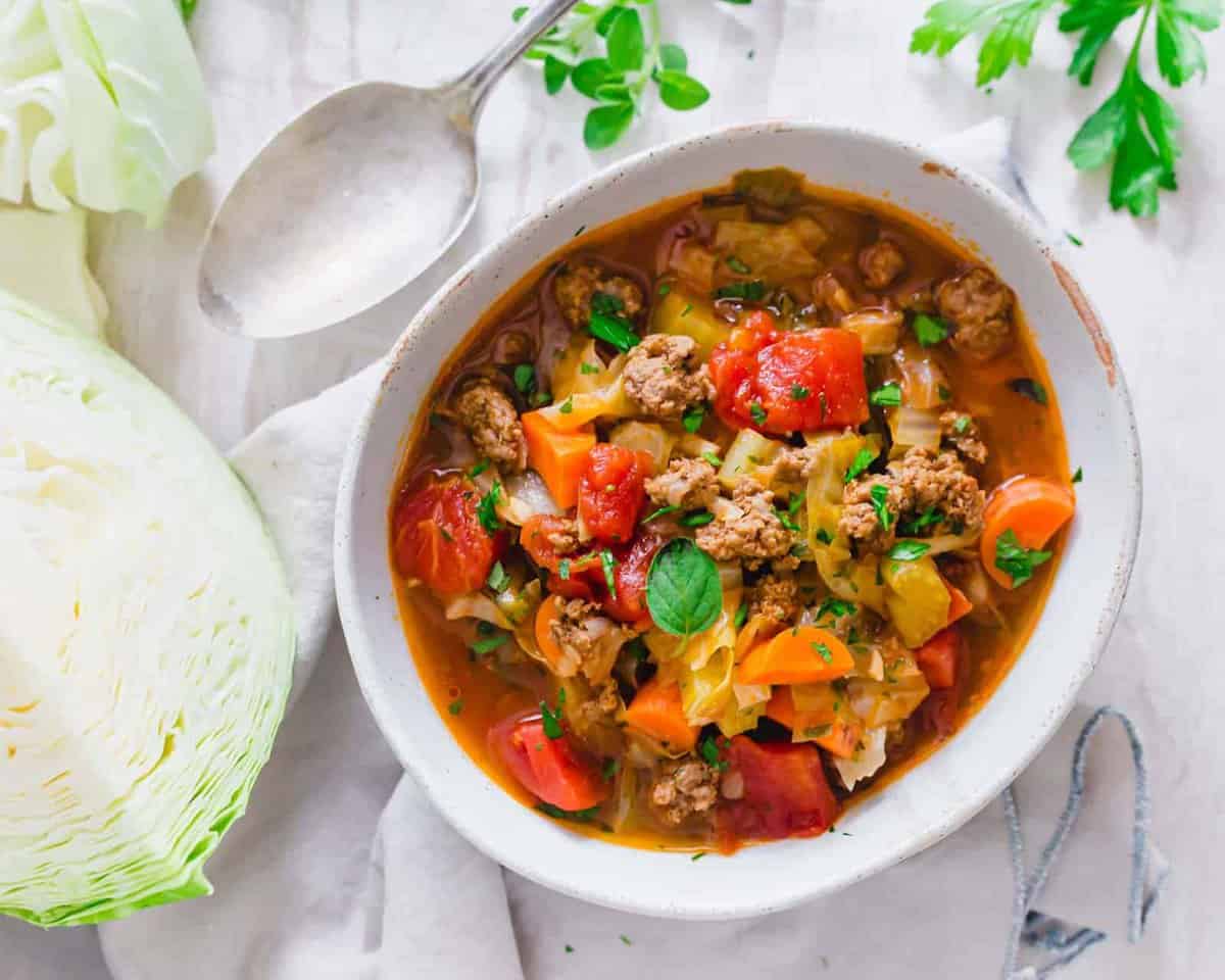 A bowl of cabbage soup with ground meat, tomatoes, and herbs, next to a spoon and a wedge of cabbage on a white surface.